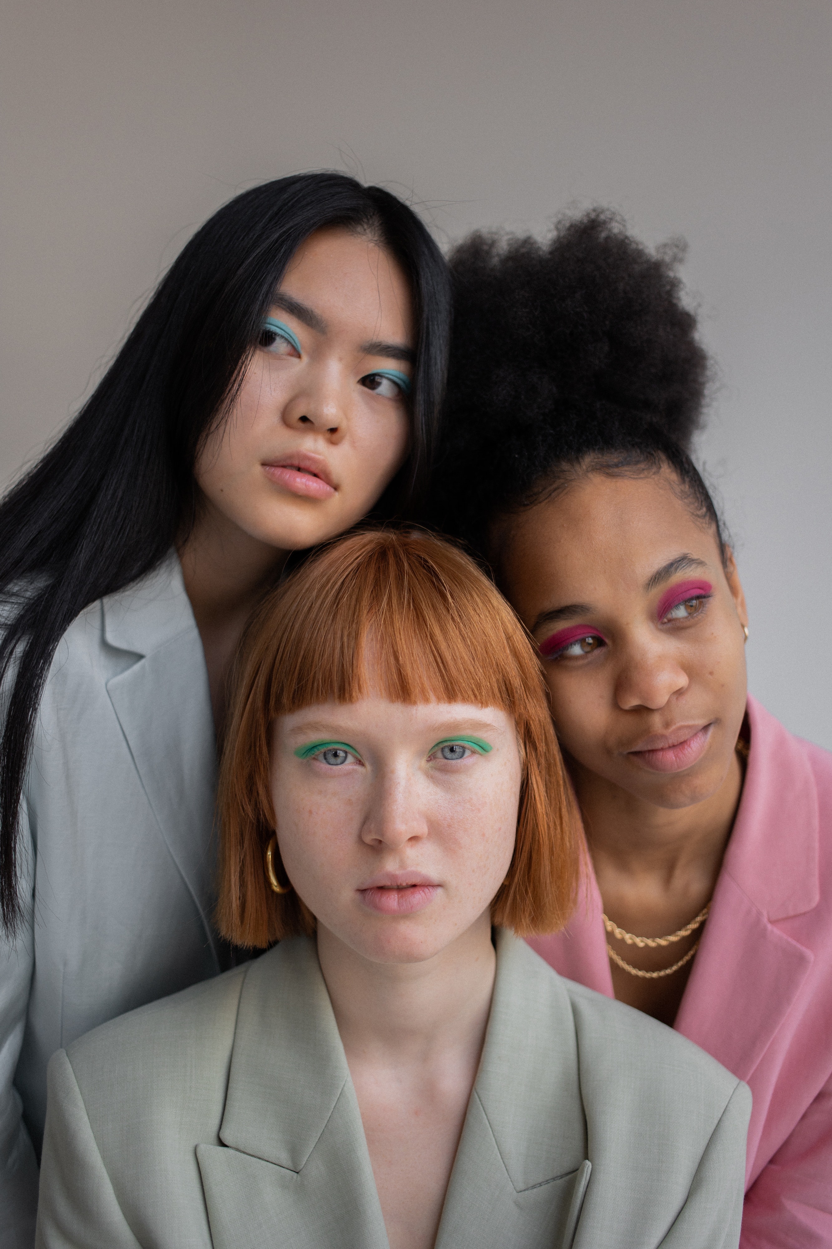 Studio photograph with an off white background and three females posing in front of the camera. Their heads are leaning 
                            against one another, with two females heads leaning against another female. The female on the left has long straight black hair and blue eyeshadow and is wearing a light
                            blue blazer. The female beside her is wearing purple and pink mixed colour eyeshadow and has brown and very curly hair, tied iup in a high ponytail. She is wearing a baby pink 
                            coloured blazer. The girl at the bottom has straight short orange brown hair with bangs. She is wearing green eyeshadow and a soft green pastel blazer.