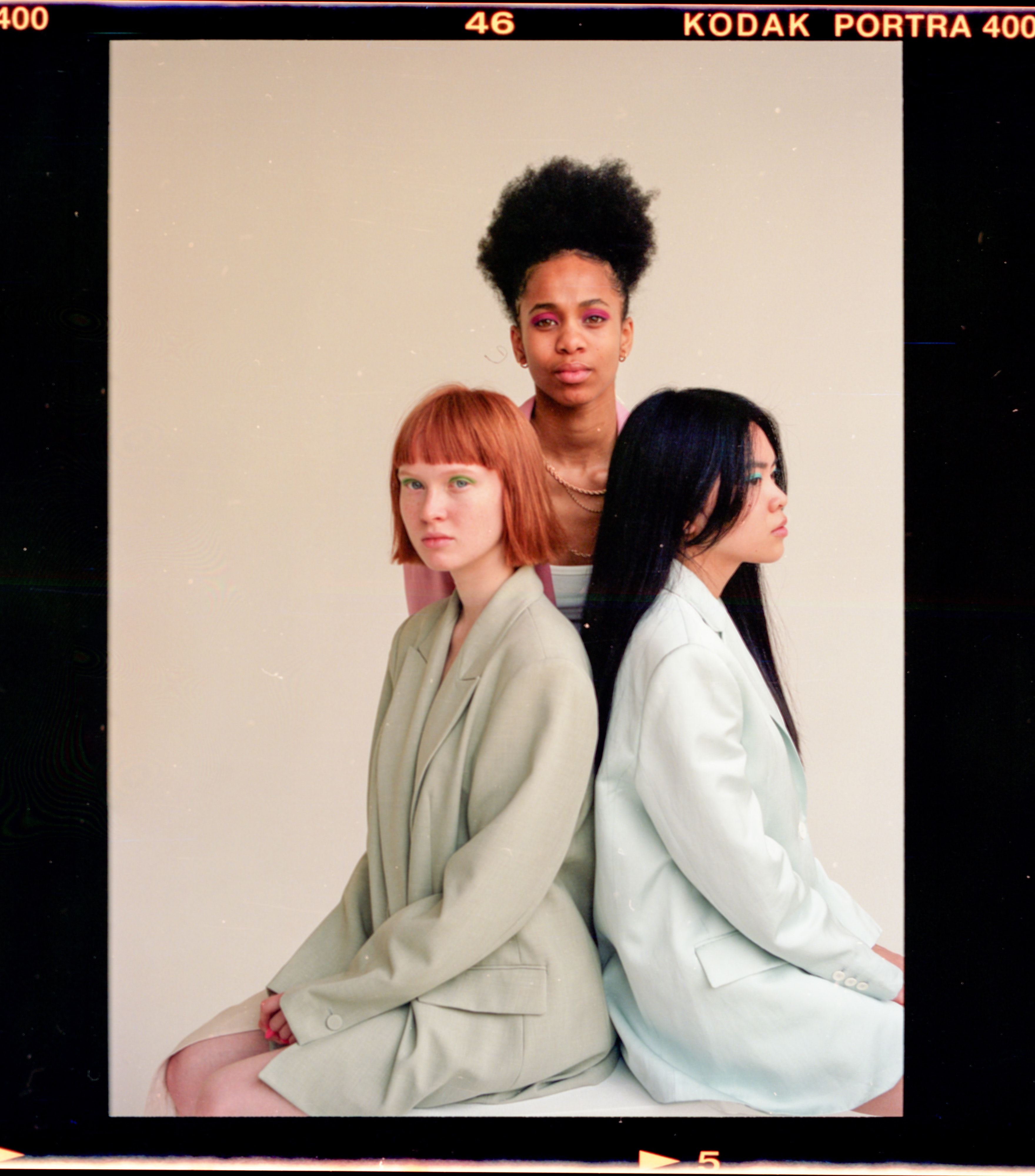 Film studio photography of three females posing for the camera. There are two females sitting down, the one on the left is facing the 
                            camera and is wearing a light green pastel blazer and her hair is straight short orange brown with bangs. The female to her left is facing away from the camera and has long black hair
                            and is wearing a blue blazer. The female in standing behind them if looking at the camera, and is wearing a white singlet and pink blazer, she also has very curly dark brown hair, tied 
                            up in a ponytail.