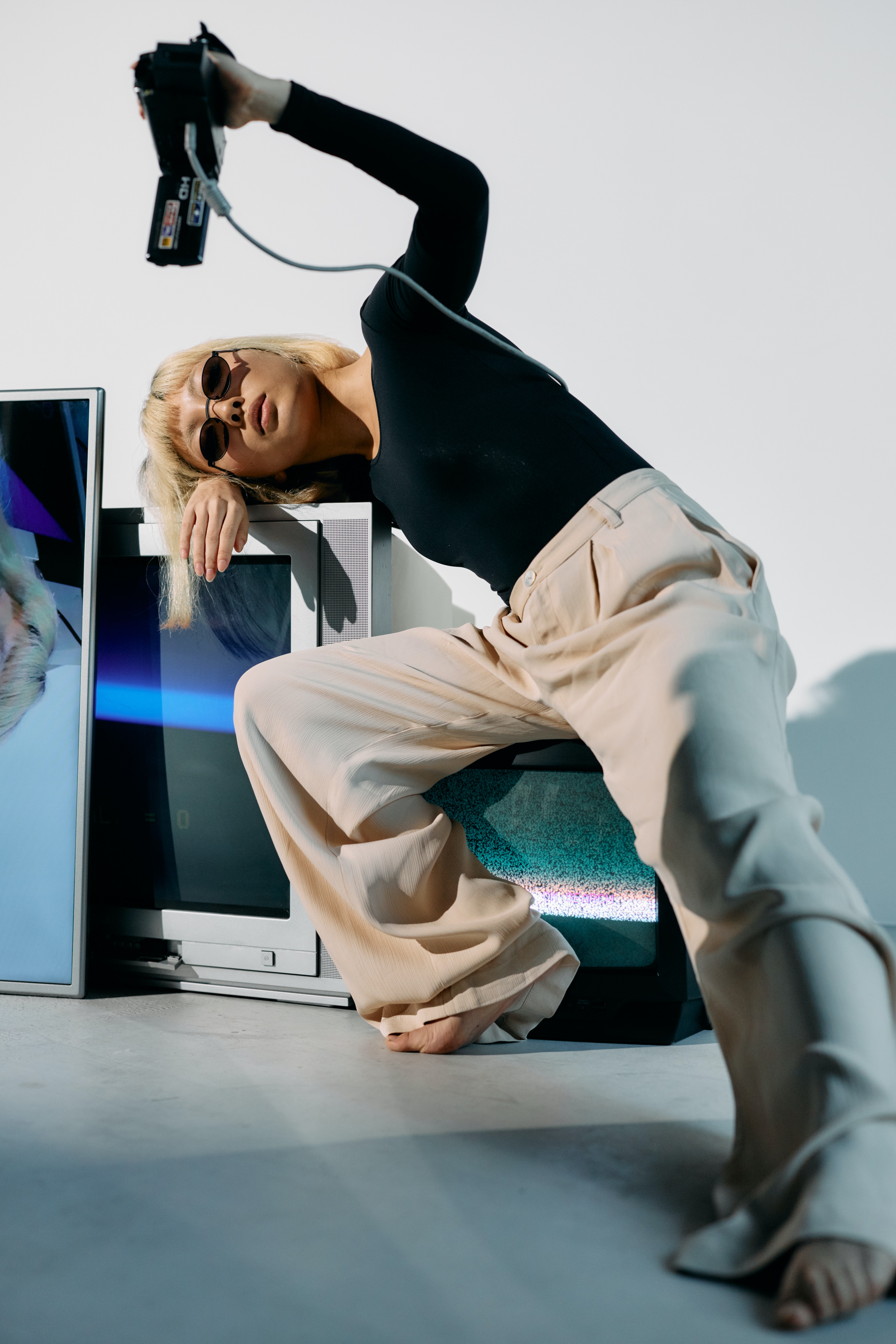 Studio photograph of a female with a short bob blonde haircut with bangs. She is wearing oval shaped sunglasses on her face and is wearing a black longsleeve
                            top with beige coloured pants. She is holding a video recorder above her and is sitting on a small black boxed shaped TV and is leaning down on a larger grey box TV, that have been placed on the 
                            floor.