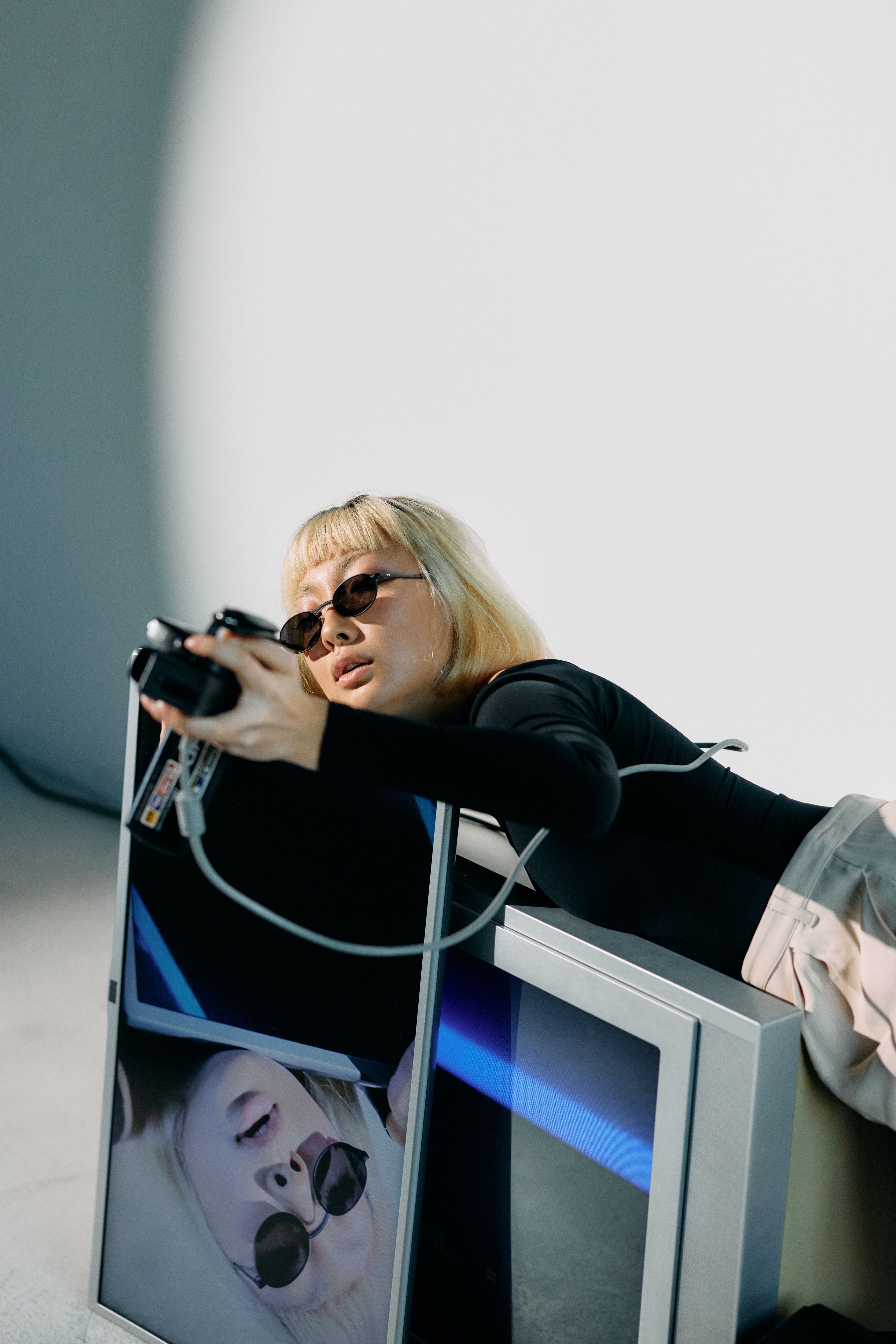 Studio photograph of a female with a short bob blonde haircut with bangs. She is wearing oval shaped sunglasses on her face and is wearing a black longsleeve
                            top with beige coloured pants. She is holding a video recorder in front of her and is lying down on a grey box TV, with her face being displayed on a slim TV, that is in front of the grey box TV.
                            The TVs have been placed on the floor.