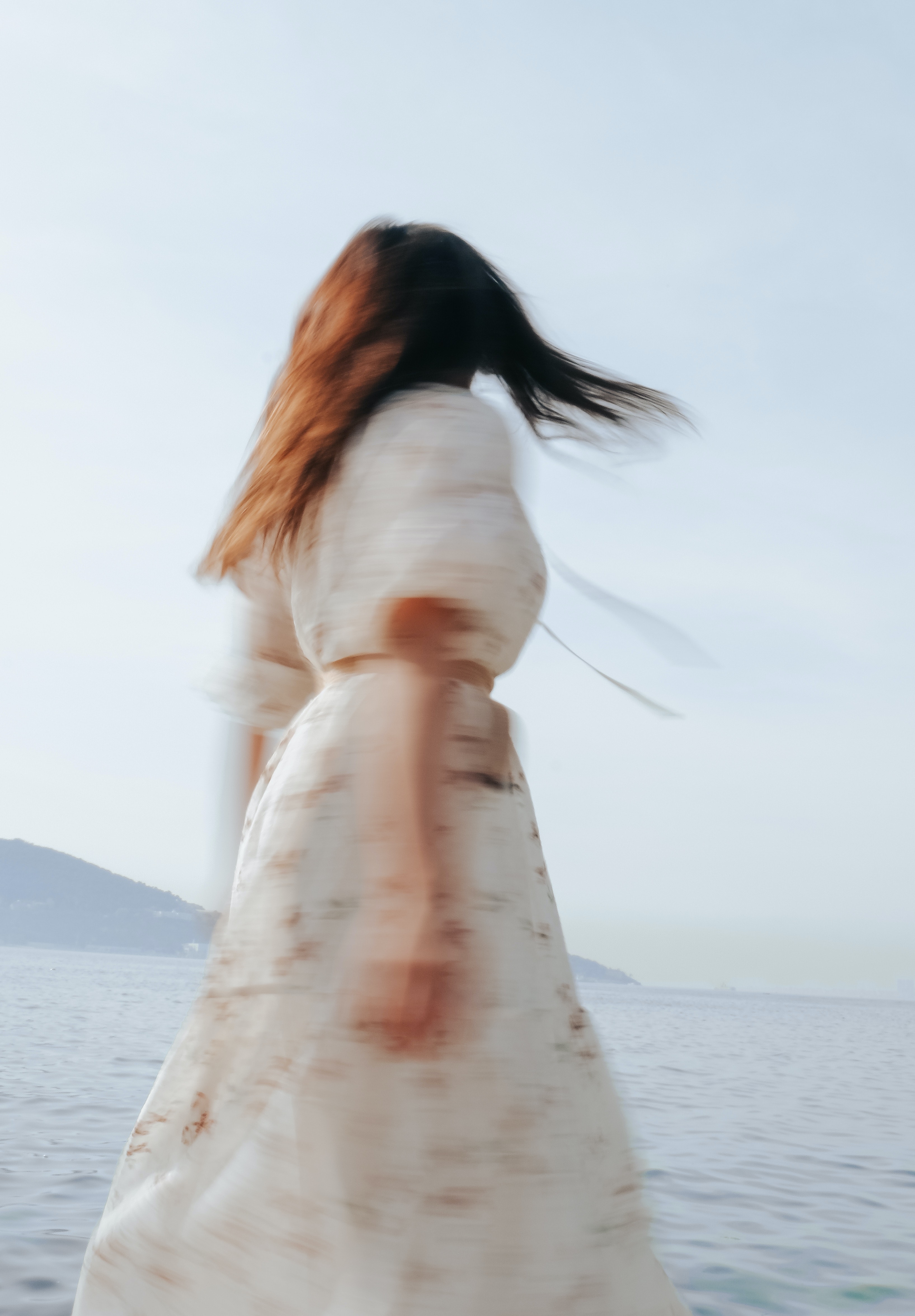 Blurry photography of a woman in a dress spinning around. She has black hair, and when the sun hits her hair it looks like a golden brown colour. 
                            Her dress is white and has a pattern on it, but can't be seen because of the blurriness of the image. The background has a foggy atmosphere, with a hill and the sea as the background.