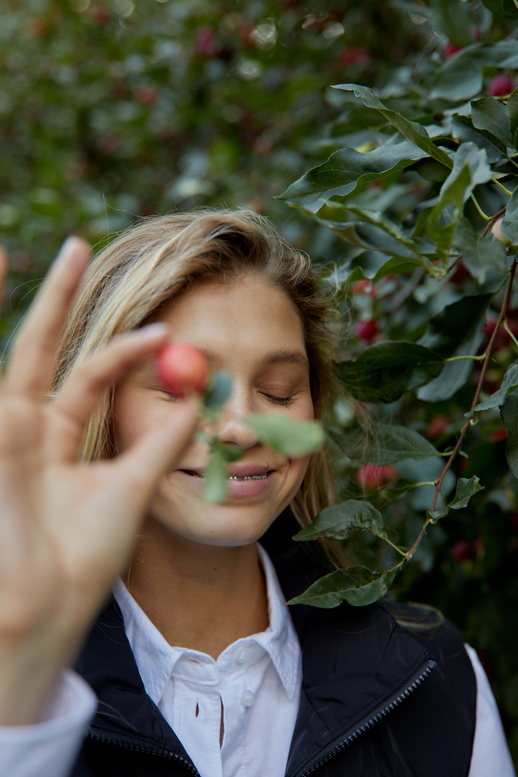 Photography of Ivy Mae holding a small pinkish red circle fruit to the camera. She has blonde hair, with brown roots
                and it is slightly curly and frizzy. The background is green, with traces of red colours from the fruit in the background. She is wearing a white button down top, with a black vest
                on top of it.