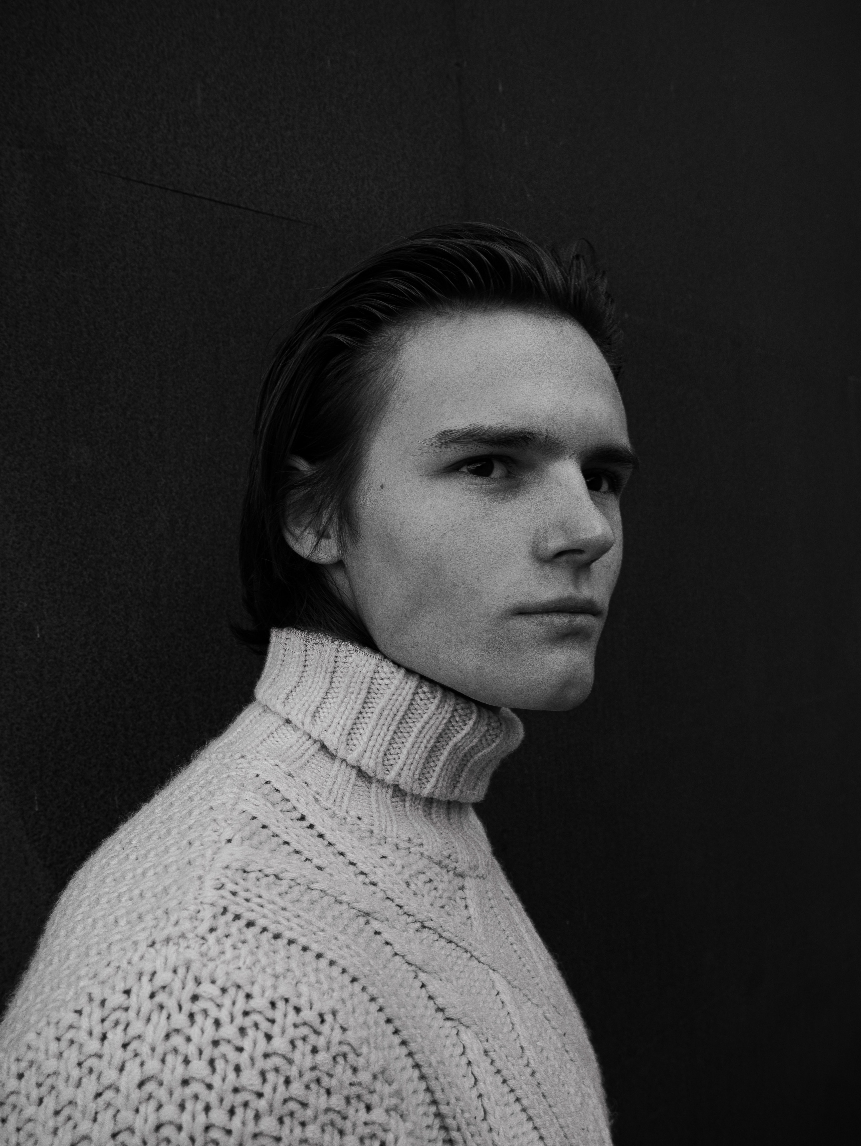 Black and white photograph of a young male in a turtleneck sweater and long slicked back hair. He has a serious face on.