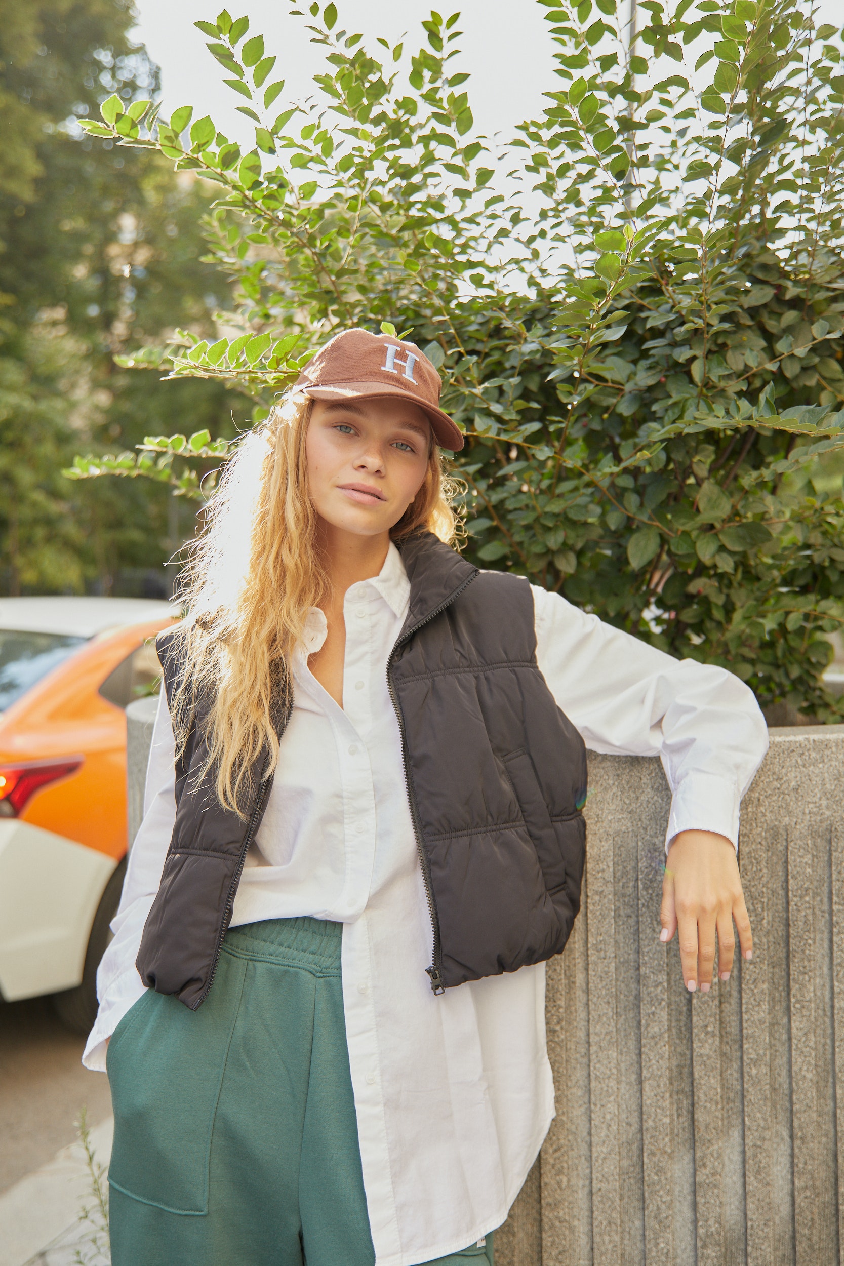 Ivy Mae wearing a brown cap with the letter H, medium length blonde wavy frizzy hair. 
                            Ivy Mae is wearing a white button down top, with a cropped vest over the top and faded moss green sweatpants. She is leaning behind a fence.