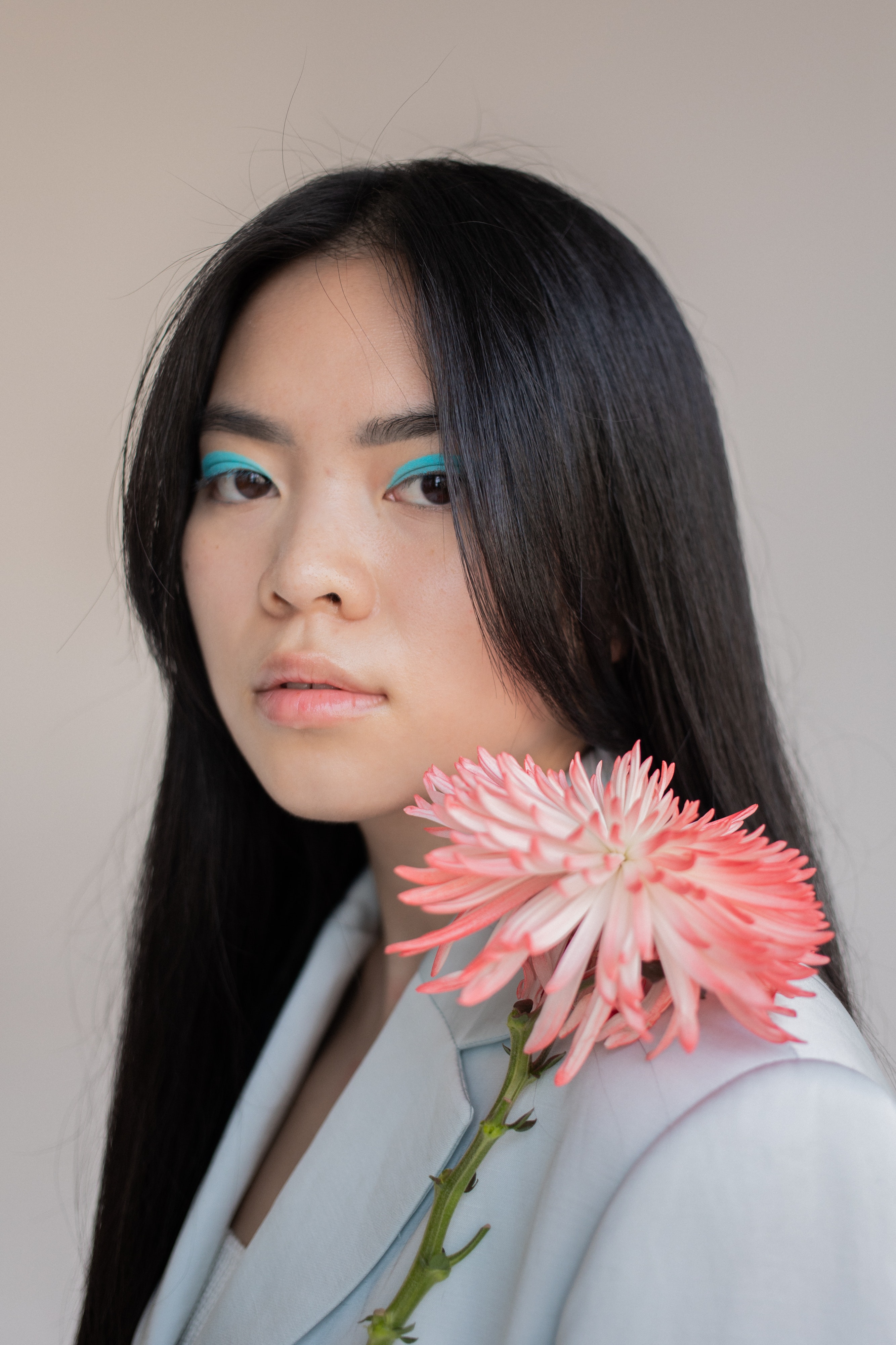 Studio portrait photograph of a female with blue eyeshadow, long black hair, who is wearing a singlet and a blue blazer over the top. She has a pink 
                            flower with lots of petals, and a green stem, sitting on her shoulder. She is sitting at angle from the camera.