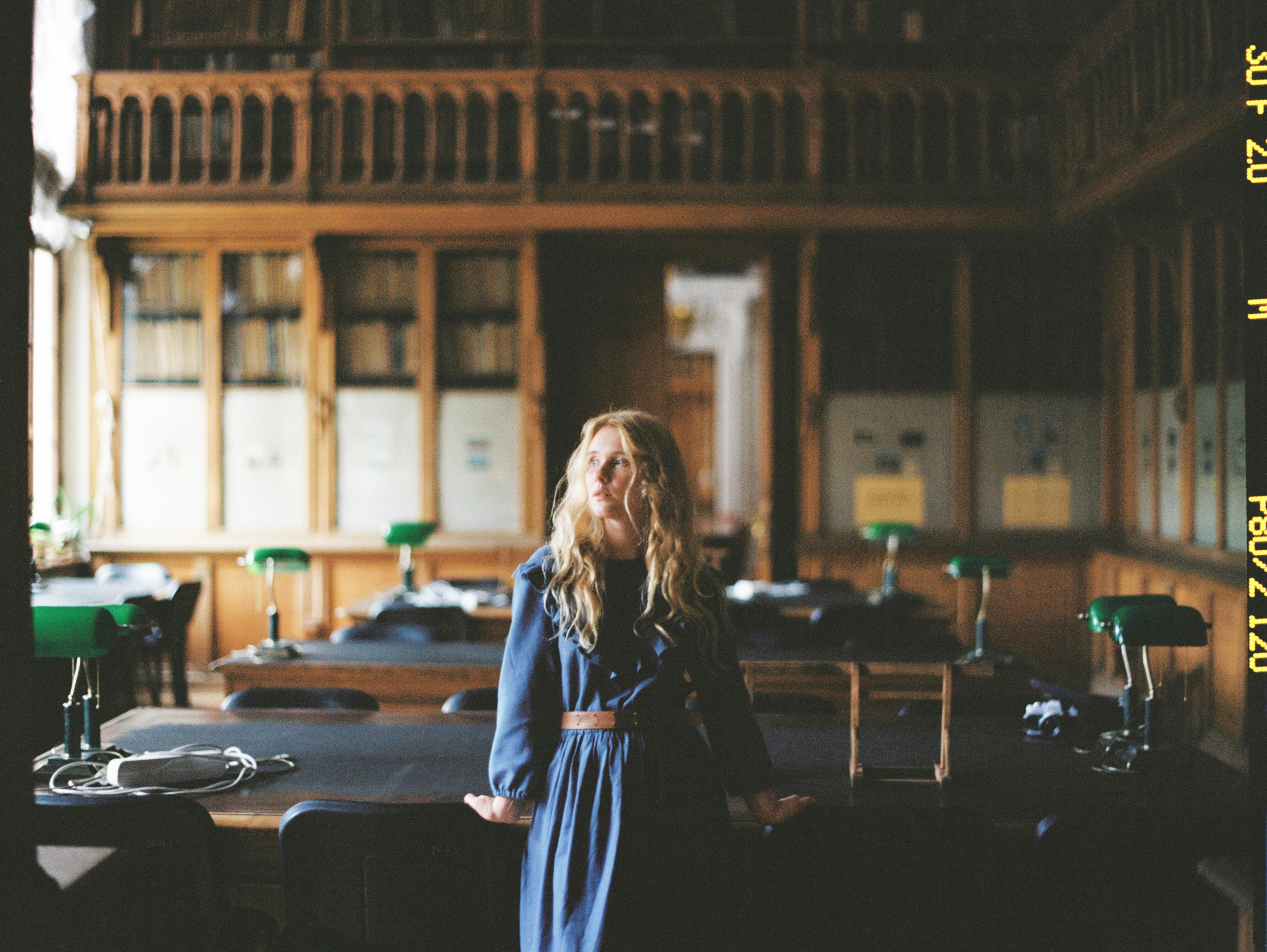 Film photograph of a woman in a long sleeve blue dress, with her hands leaning on chairs behind her, in a library.