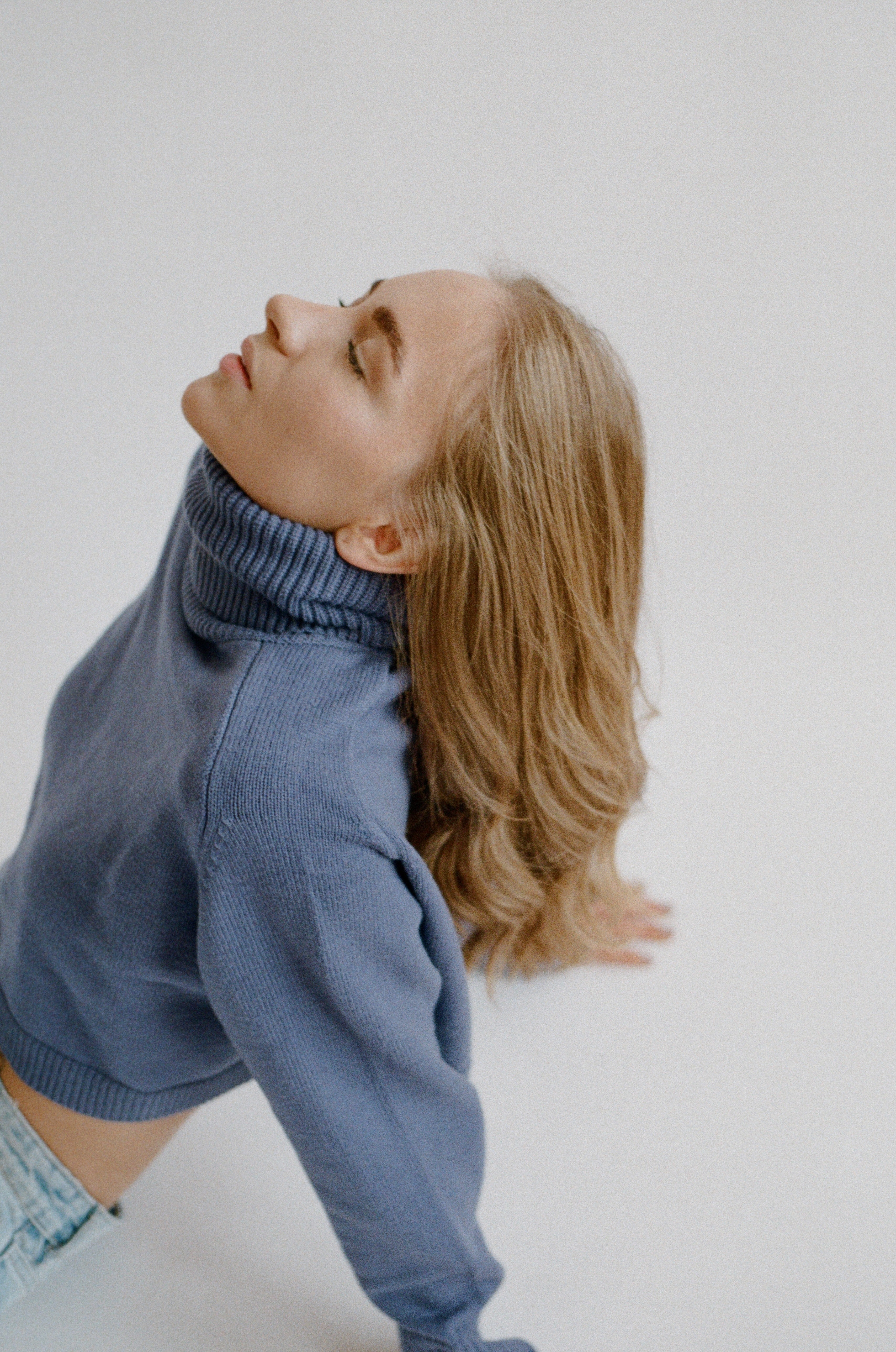 Bright studio photograph with a white backdrop, a woman in a cropped blue sweater posing on the floor with hands outstreched behind her, eyes closed and facing up towards the ceiling.