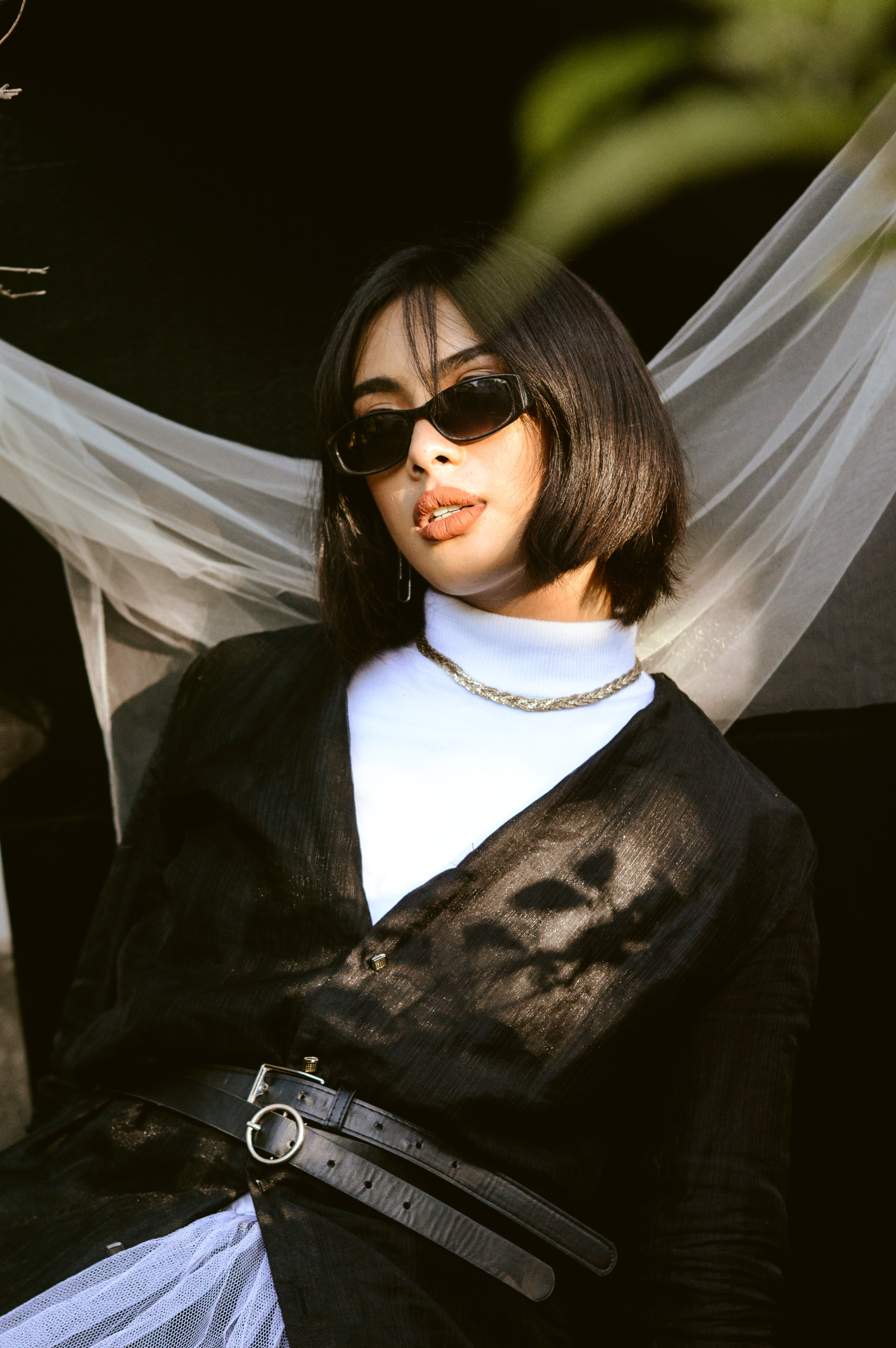 Image of a female with a black hair, styled as a short bob with bangs. She is wearing light makeup and sunglasses on her face, and is wearing
                            a white turtleneck and black blazer, tied with two belts at her waist. She has a gold necklace around her neck. She is leaning behind something and there is a white see through fabric behind
                            her. There is a green leaf in the top right of the image and it is blurred out.