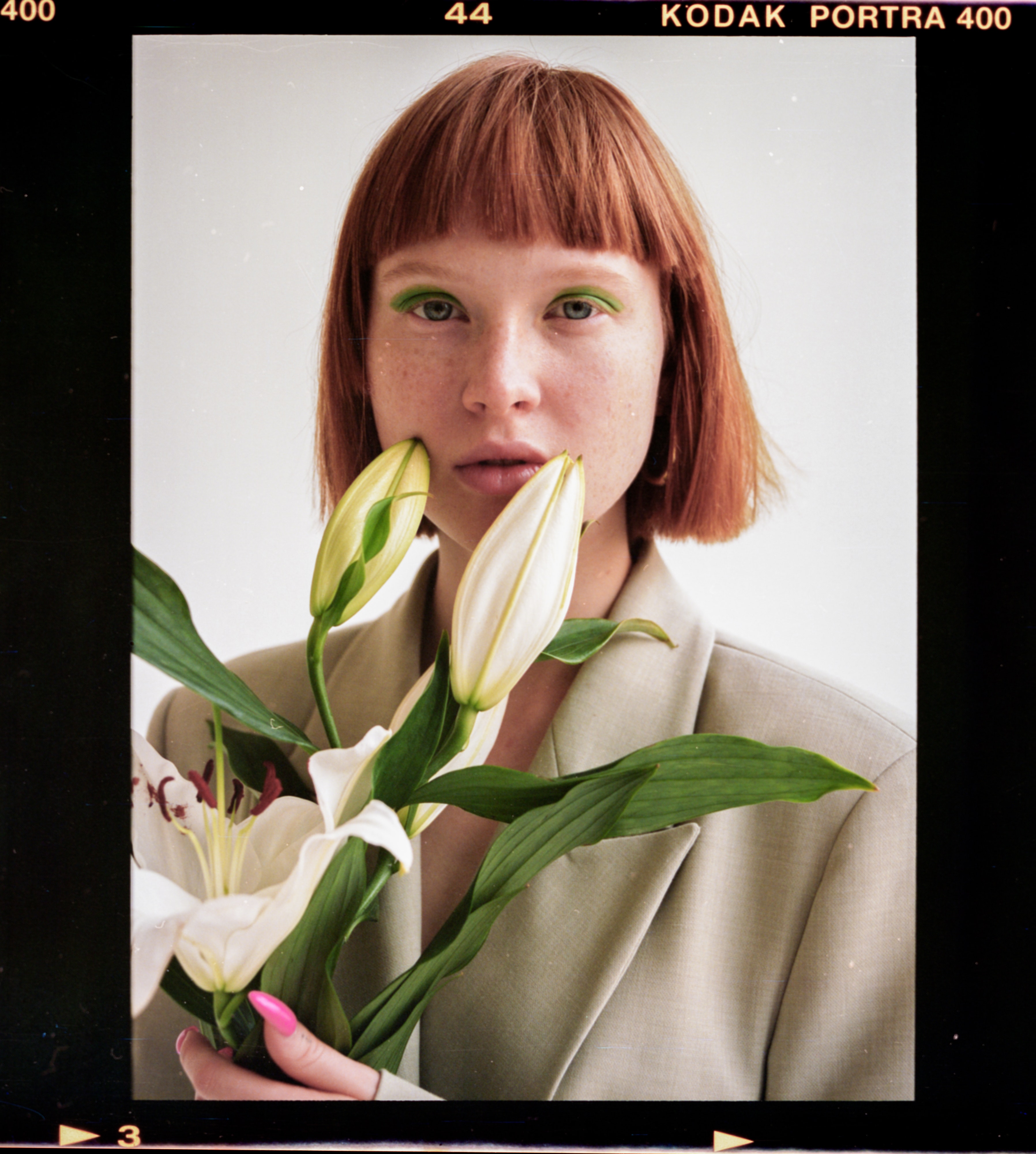 Young woman with orange brown hair, green eyeshadow and freckles holding flowers near her face.