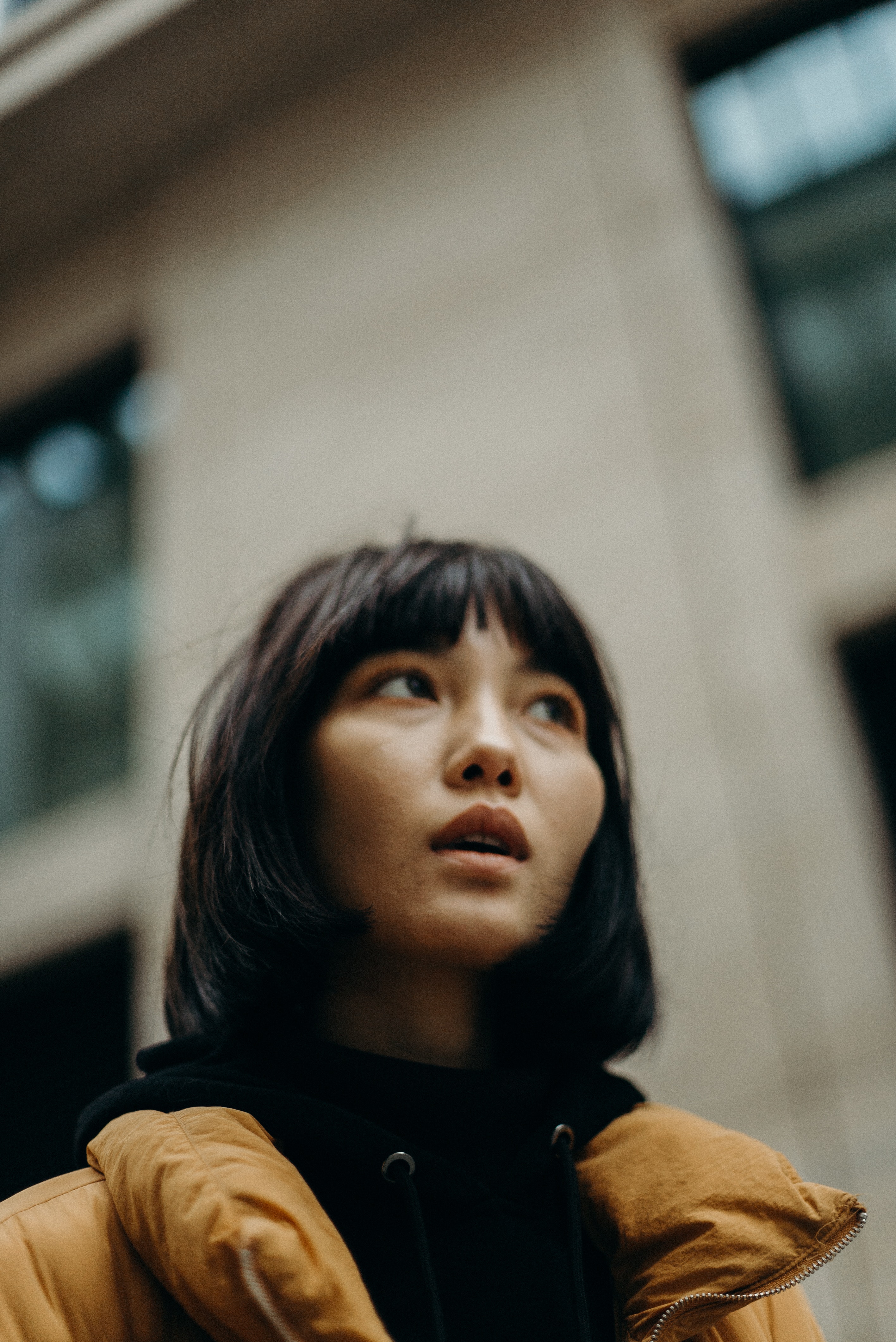 Photo of a young woman with black hair and wearing a yellow jacket and black hoodie, taken at a low angle in front of a building.